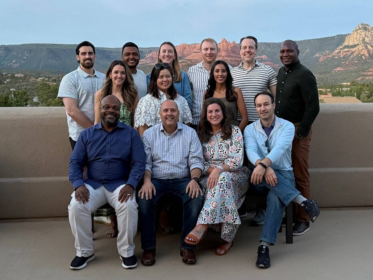 13 coworkers pose together on a rooftop terrace at dusk, with red rock desert mountains in the background during a team retreat.