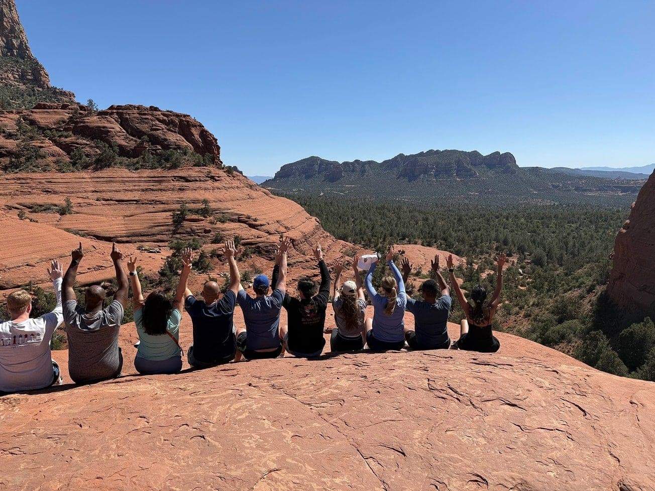 A group sits on a red rock ledge in Sedona with arms raised, overlooking a forested valley and distant buttes.